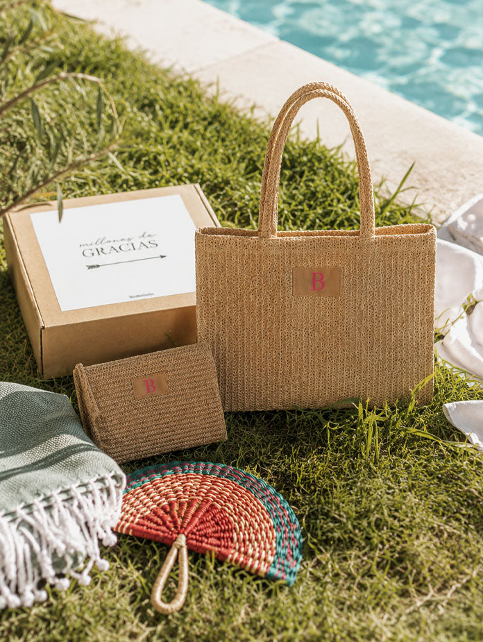 Bolso de yute personalizado con inicial bordada y cartera a juego, presentado junto a un abanico artesanal y una caja de regalo junto a una piscina.